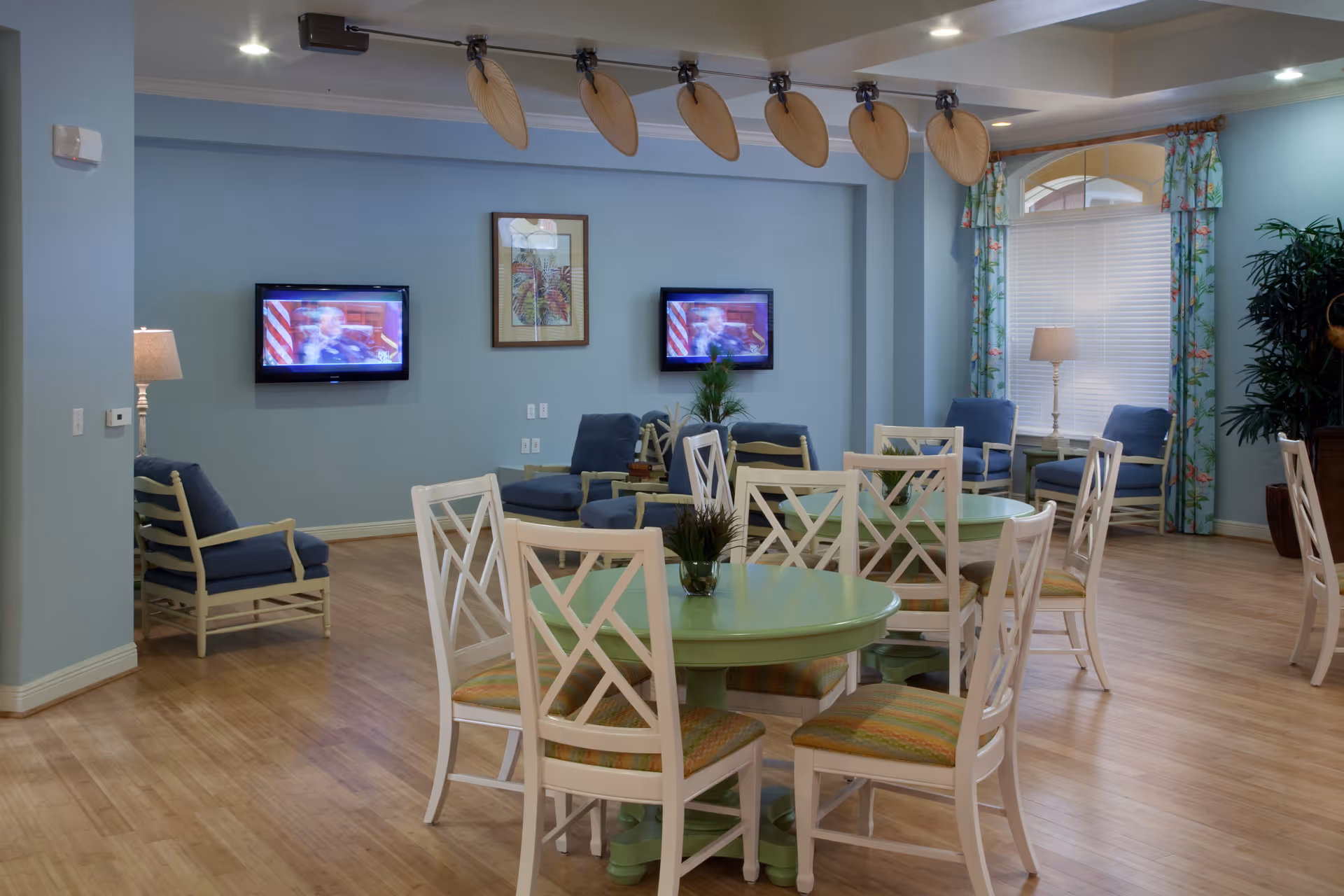 A spacious common area with light blue walls and wooden flooring featuring round green tables surrounded by white chairs with patterned cushions. There are two flat-screen TVs mounted on the wall, a framed picture between them, and several blue cushioned armchairs arranged around the room. The room is well-lit with ceiling lights and lamps, and there are floral curtains on the windows along with a large potted plant in the corner.