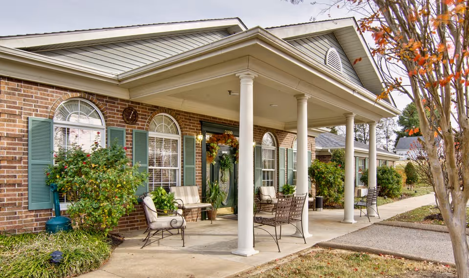 Brick-front senior living entrance with white columns, outdoor seating, green shutters, and landscaped shrubs along a covered porch.
