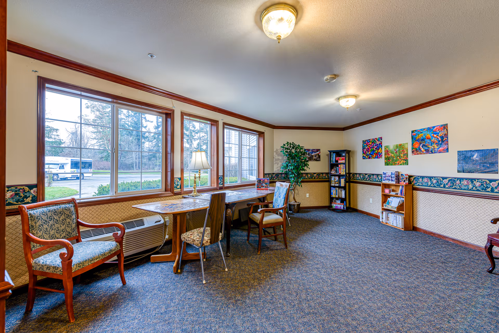 Sunlit common room with tables, chairs, bookshelves, artwork on the walls, and large windows looking outside.