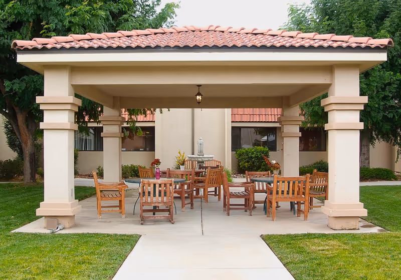 Covered outdoor patio with wooden tables and chairs under a tiled roof in front of a building.