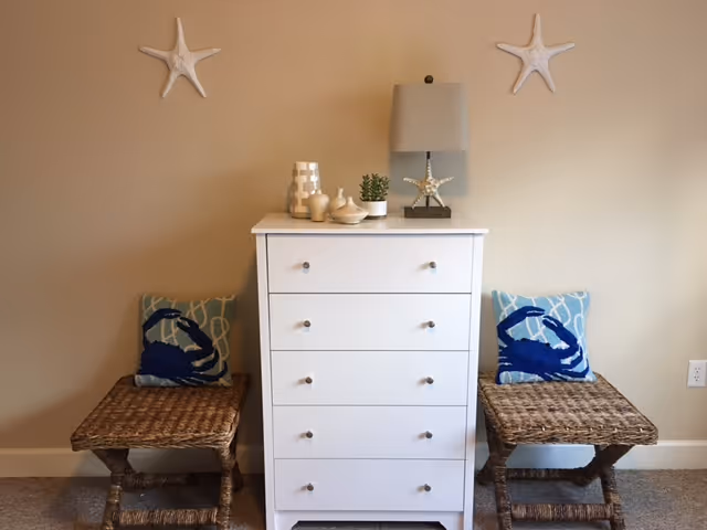 A white chest of drawers with decorative items on top including a lamp with a starfish base, a small plant, and vases. On either side of the chest are two wicker stools with blue crab-themed cushions. Two white starfish wall decorations are mounted above the chest on a beige wall.