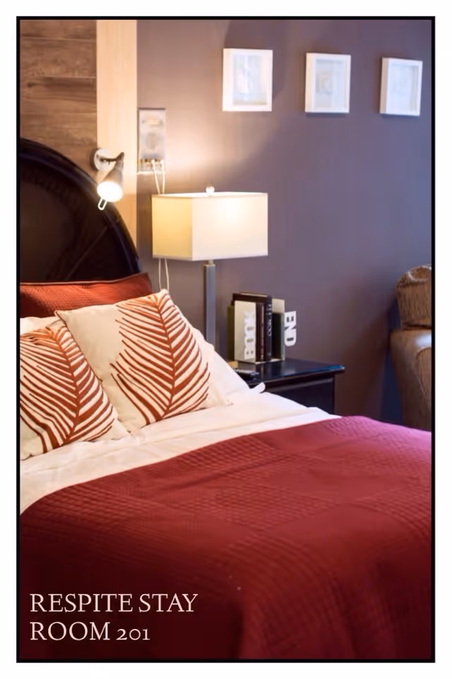 A cozy bedroom with a bed featuring a red quilt and decorative pillows with a leaf pattern. There is a bedside table with books and a modern lamp, and three framed pictures hang on the wall above the bed. A small reading light is attached to the headboard.