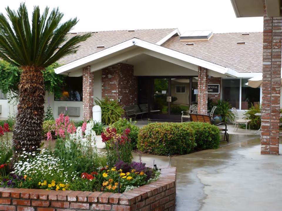 Brick-front retirement community entrance with a covered porch, benches and colorful flower beds in front.