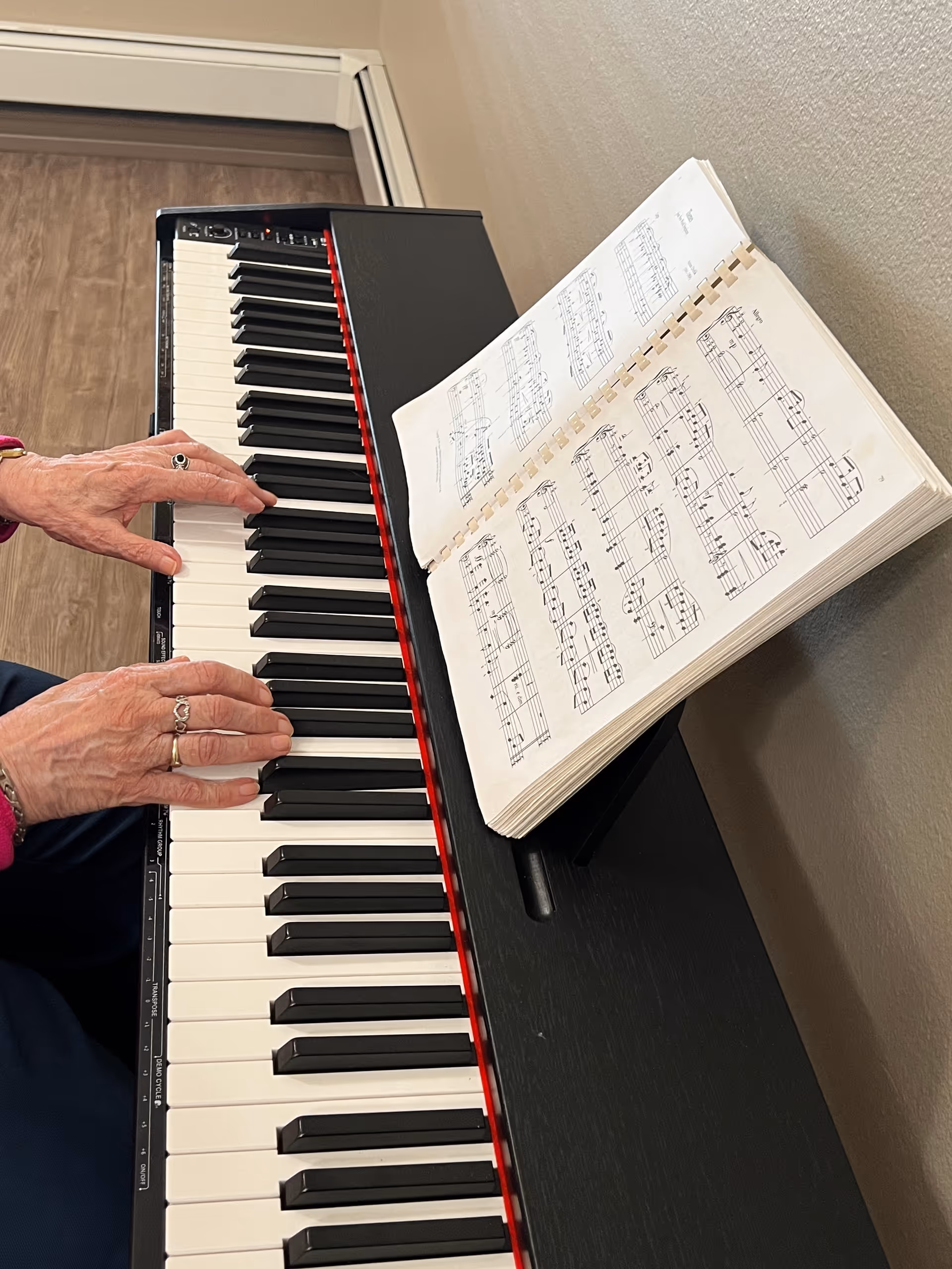 Close-up of an elderly person's hands playing a digital piano with sheet music placed on the piano stand, against a beige wall and wooden floor background.