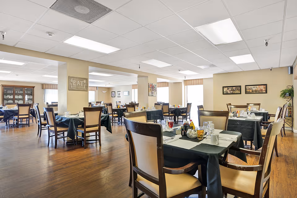 A spacious dining room with multiple tables covered in dark tablecloths, each set with glasses, cups, napkins, and condiments. The room has wooden flooring, beige walls, and large windows letting in natural light. There are framed pictures on the walls and a cabinet with dishes in the background.
