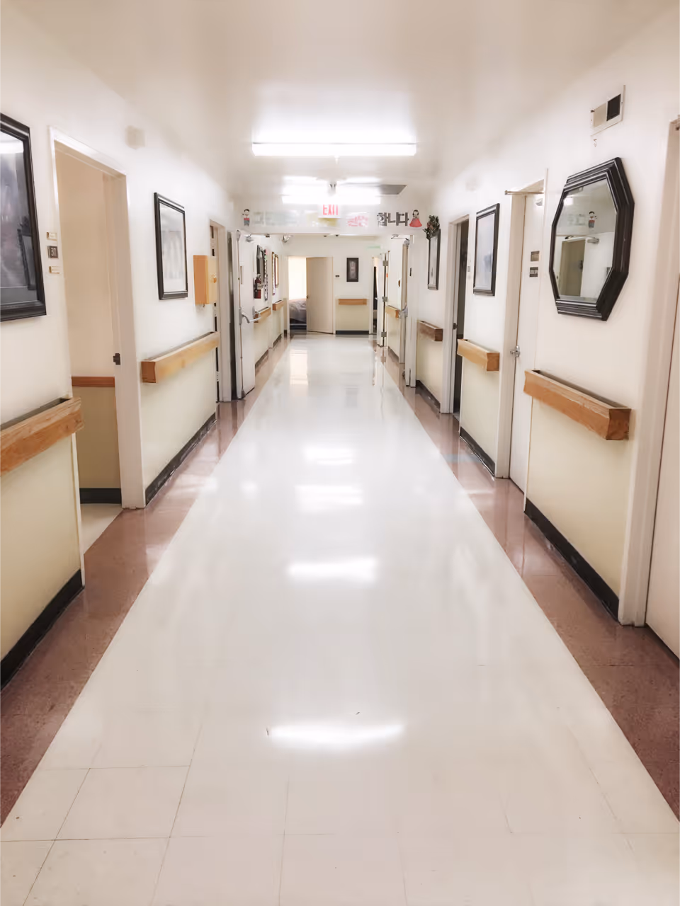 A long, clean hallway in a care facility with white walls and a shiny white floor. The hallway has several doors on both sides, wooden handrails along the walls, framed pictures, and a mirror. Bright fluorescent lights illuminate the corridor, and an exit sign is visible at the far end.
