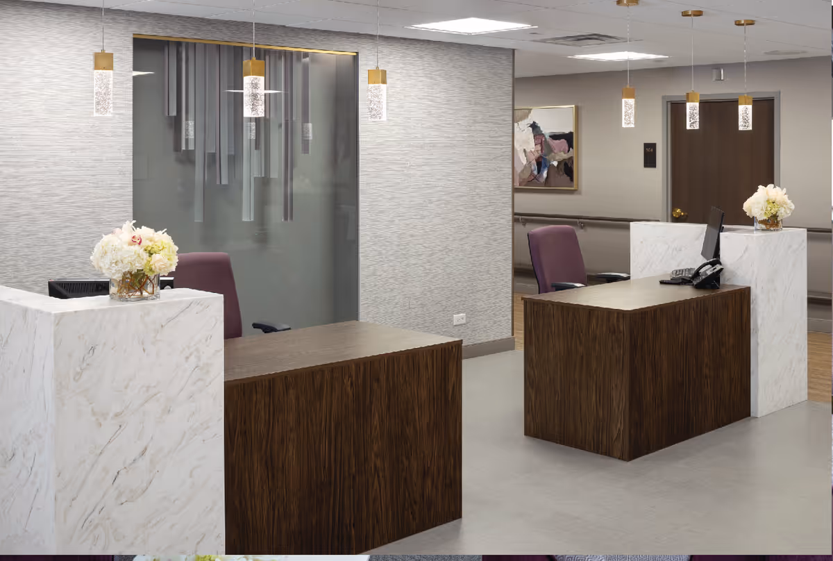Reception area with two wooden desks featuring white marble accents, each with a computer and a vase of white flowers. Behind the desks are purple office chairs, pendant lights hanging from the ceiling, and a hallway with a door and framed artwork on the wall.