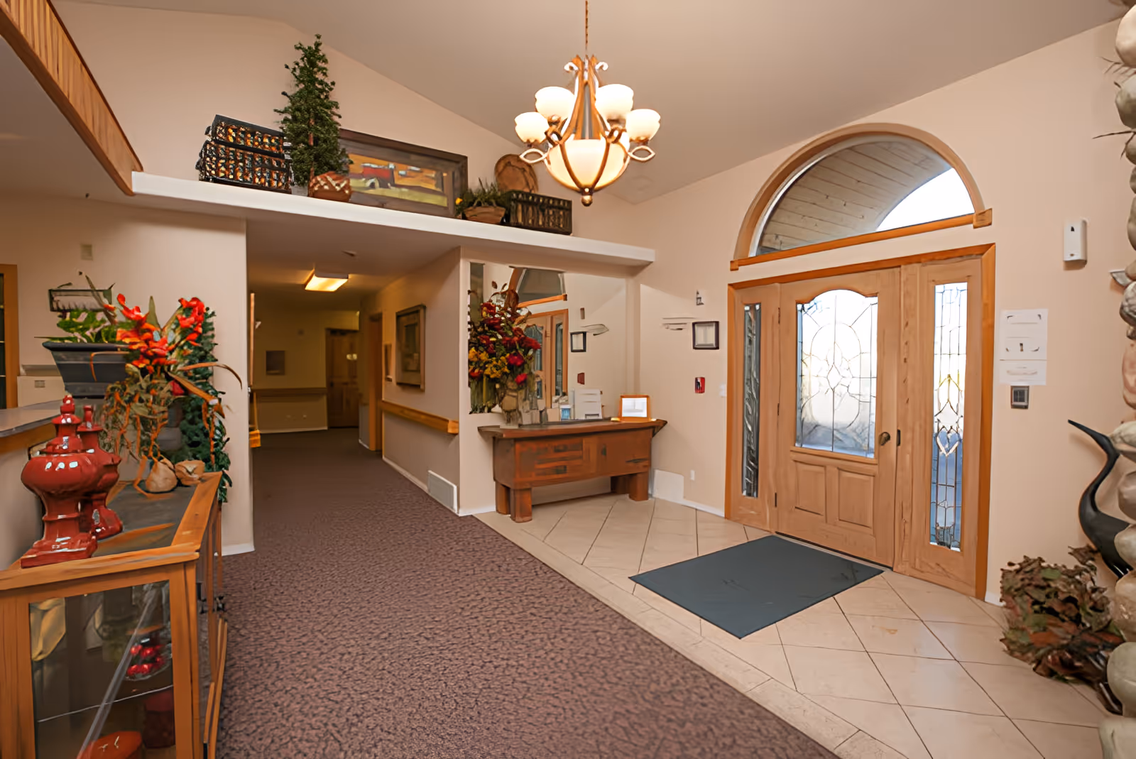 Assisted living facility lobby with double wooden entry doors, chandelier, console table and decorative plants.