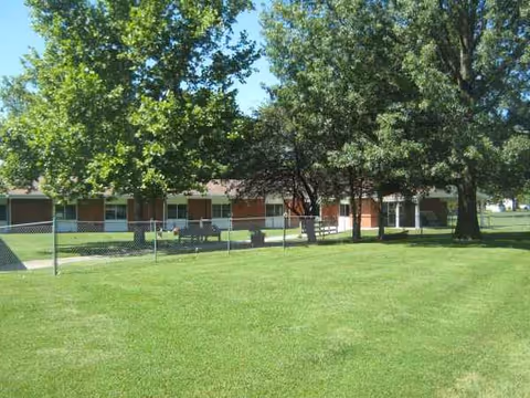 Exterior view of a single-story nursing home with a grassy lawn, trees, and a chain-link fence.