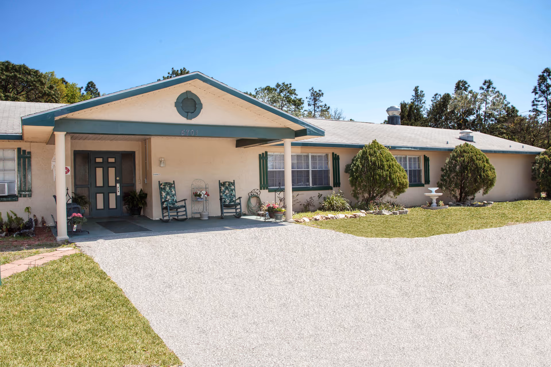 Single-story residential building with a covered front porch, rocking chairs, driveway and landscaped lawn.