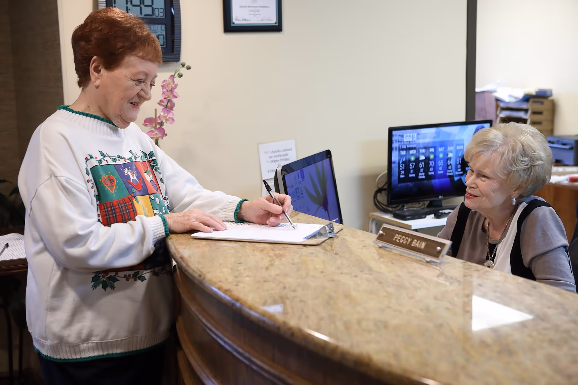 An elderly woman in a festive sweater is signing a document on a reception desk while another elderly woman, identified as Peggy Bain by a nameplate, smiles and looks on from behind the desk. A computer monitor and a digital clock are visible in the background.