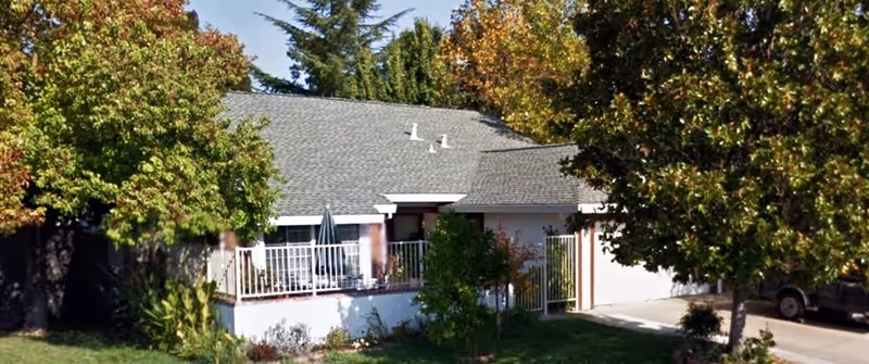 Single-story house with a covered front porch/deck, driveway, and surrounding trees.