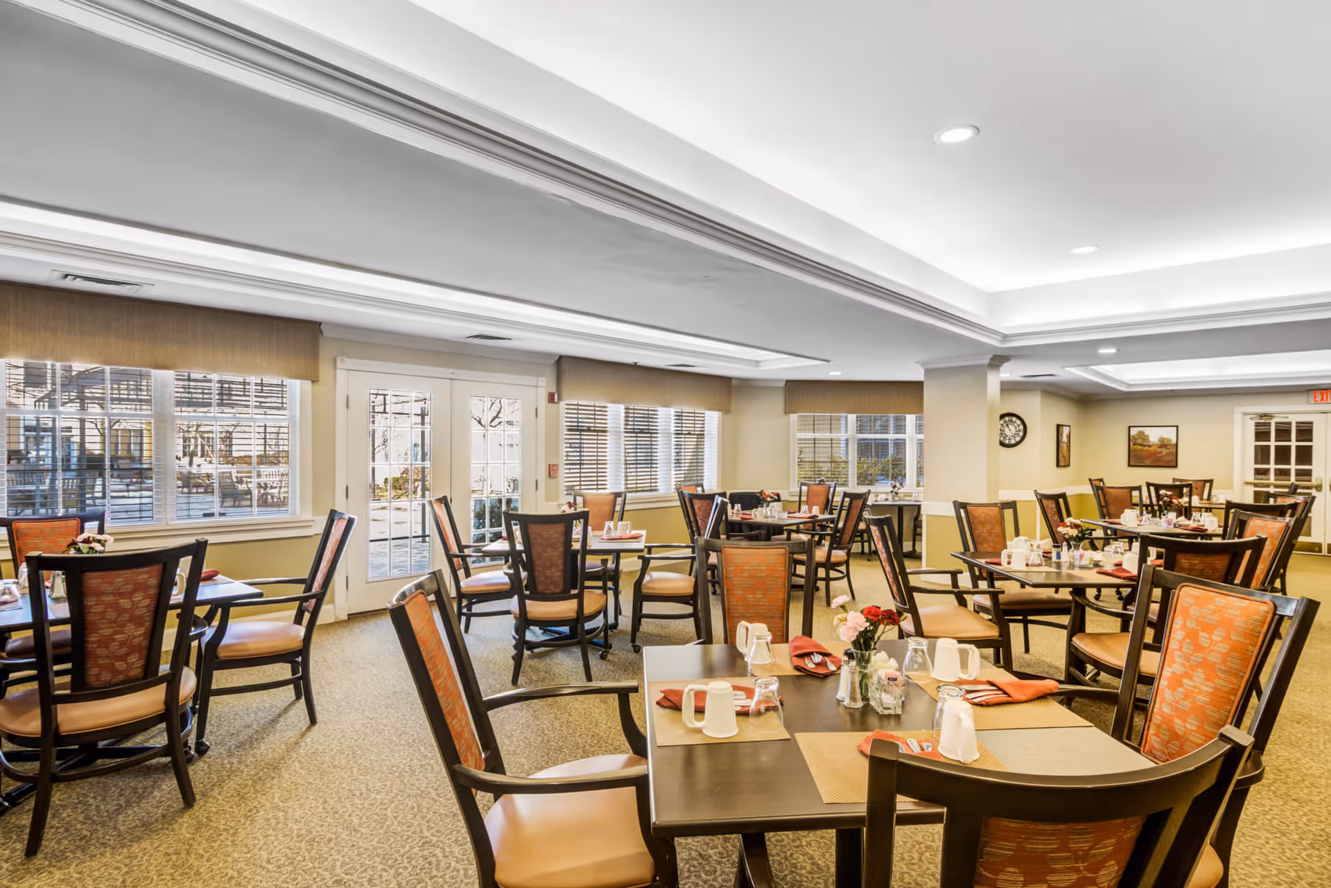 A bright and spacious dining room with multiple tables and chairs arranged neatly. Each table is set with placemats, napkins, cups, and small flower vases. Large windows and glass doors allow natural light to fill the room, and the walls are decorated with a clock and framed pictures.
