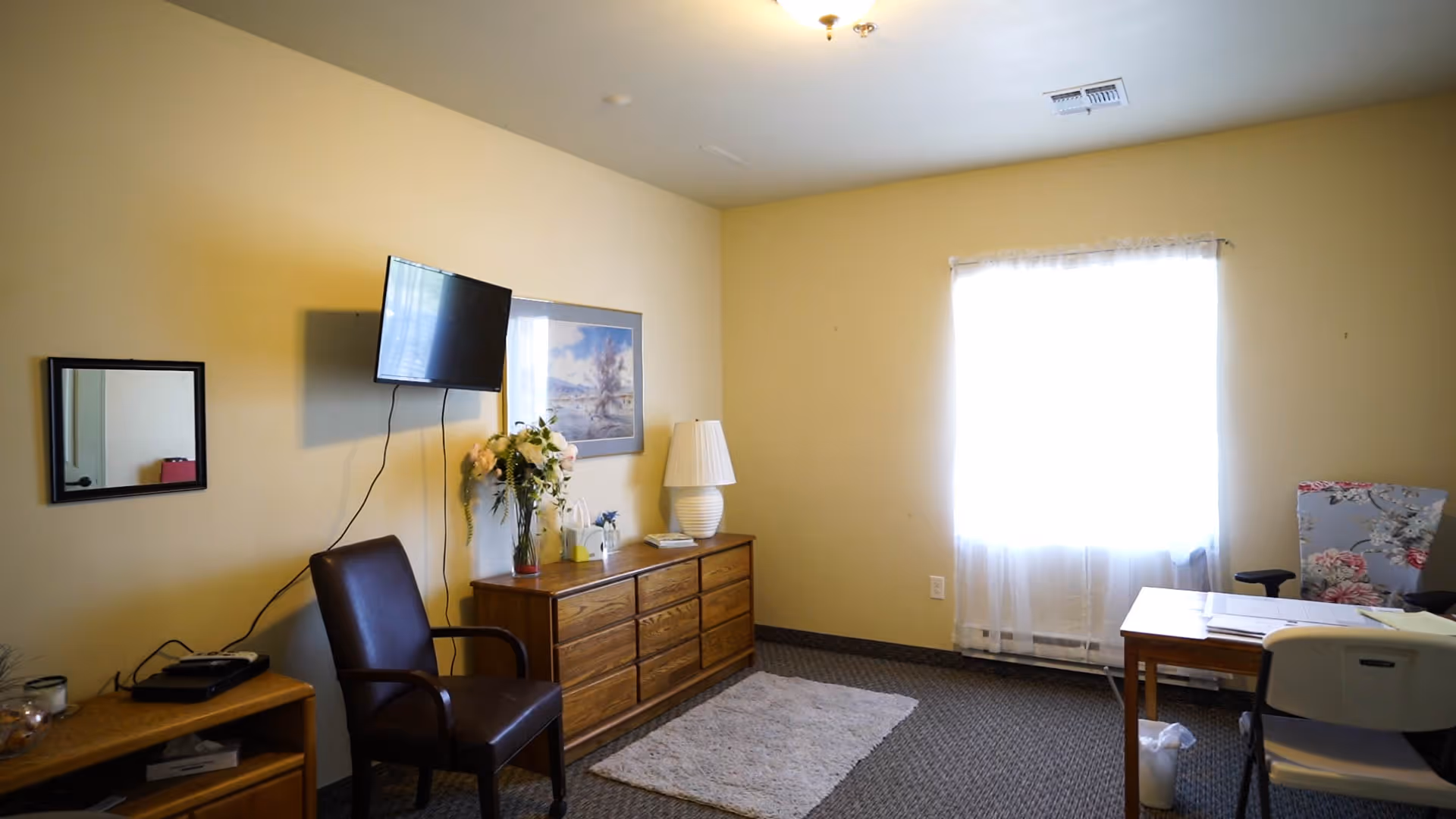 Sunny sitting room with a dresser, wall-mounted TV, chairs, a small desk, and a window with sheer curtains.