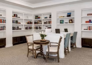 A bright and spacious library or reading room with white built-in bookshelves filled with books along the walls. There is a round wooden table with four chairs in the foreground and a long white desk with three upholstered chairs in the background. The room has a beige carpet and white walls with crown molding.