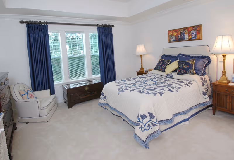 A bedroom with a large bed covered in a white and blue patterned quilt and multiple pillows. There are two bedside tables with lamps on each side of the bed. A cushioned armchair with a decorative pillow is placed near a window with blue curtains. A wooden chest is positioned under the window, and a colorful painting hangs above the bed.