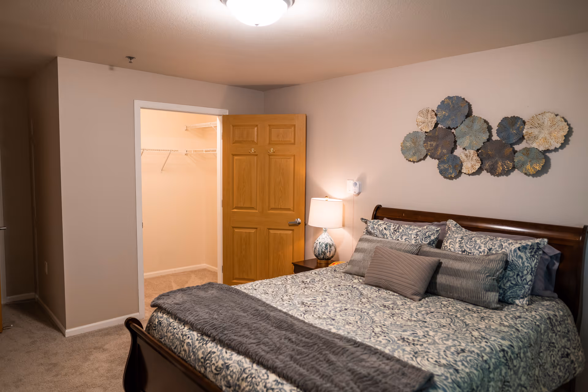 A bedroom with a large bed covered in patterned bedding and multiple pillows. A wooden nightstand with a lamp is next to the bed. The wall above the bed has decorative metal wall art. An open wooden door reveals a walk-in closet with wire shelving. The room has beige walls and carpeted floor.