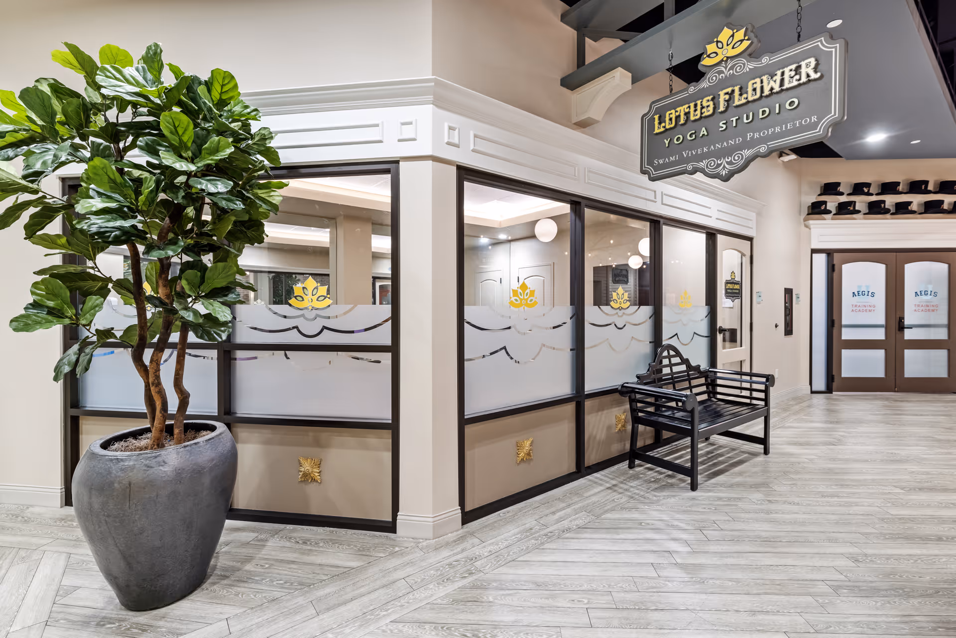 Interior view of a yoga studio named Lotus Flower Yoga Studio inside a senior living facility. The studio has large glass windows with frosted lotus flower designs, a black bench outside, a large potted plant to the left, and double doors labeled Aegis Training Academy in the background.