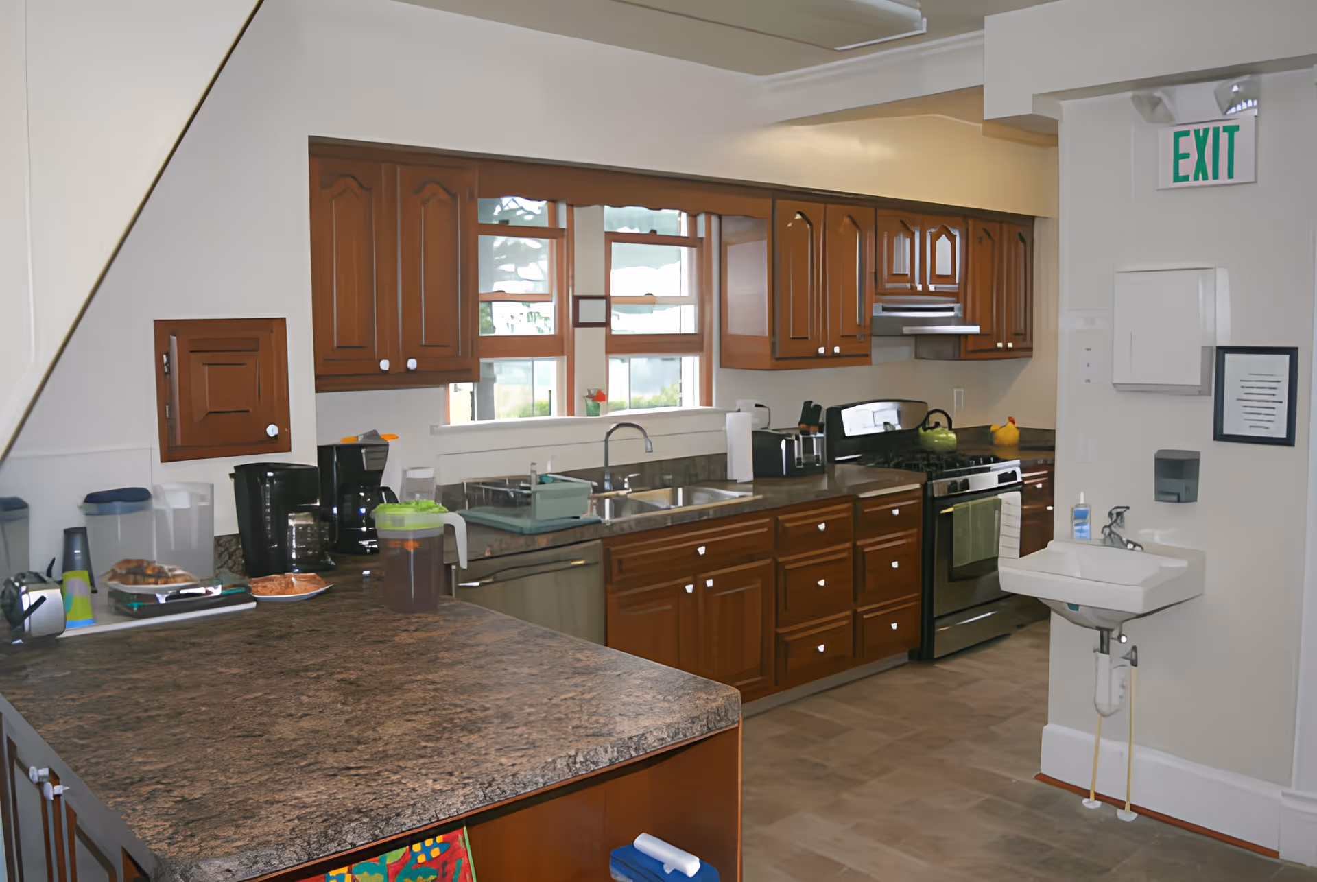 A spacious kitchen with wooden cabinets, a granite countertop island, and stainless steel appliances including a stove and dishwasher. There are two coffee makers, a pitcher of iced tea, and some baked goods on the counter. A small white sink with soap dispenser and paper towel holder is mounted on the wall near an exit sign. Large windows above the sink let in natural light.