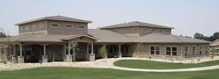 Exterior view of a single-story senior living facility building with beige walls, multiple windows, and a covered porch area, surrounded by a well-maintained lawn and a paved walkway.