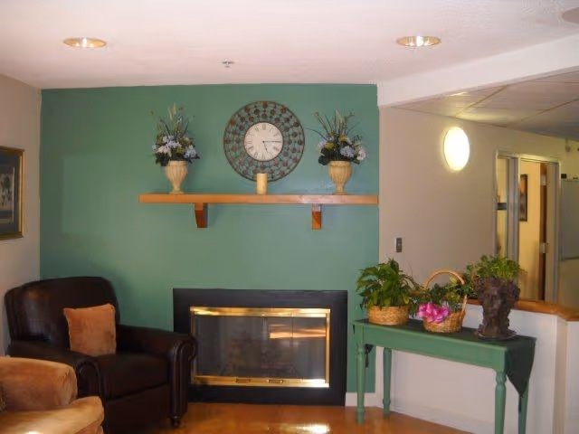 A cozy living room area with a dark brown leather armchair and a tan armchair, both with orange cushions. A green accent wall features a wooden shelf holding two flower arrangements and a clock in the center. Below the shelf is a fireplace with a glass and brass frame. To the right, a green table holds three plant arrangements. The room has wooden flooring and a hallway is visible in the background.
