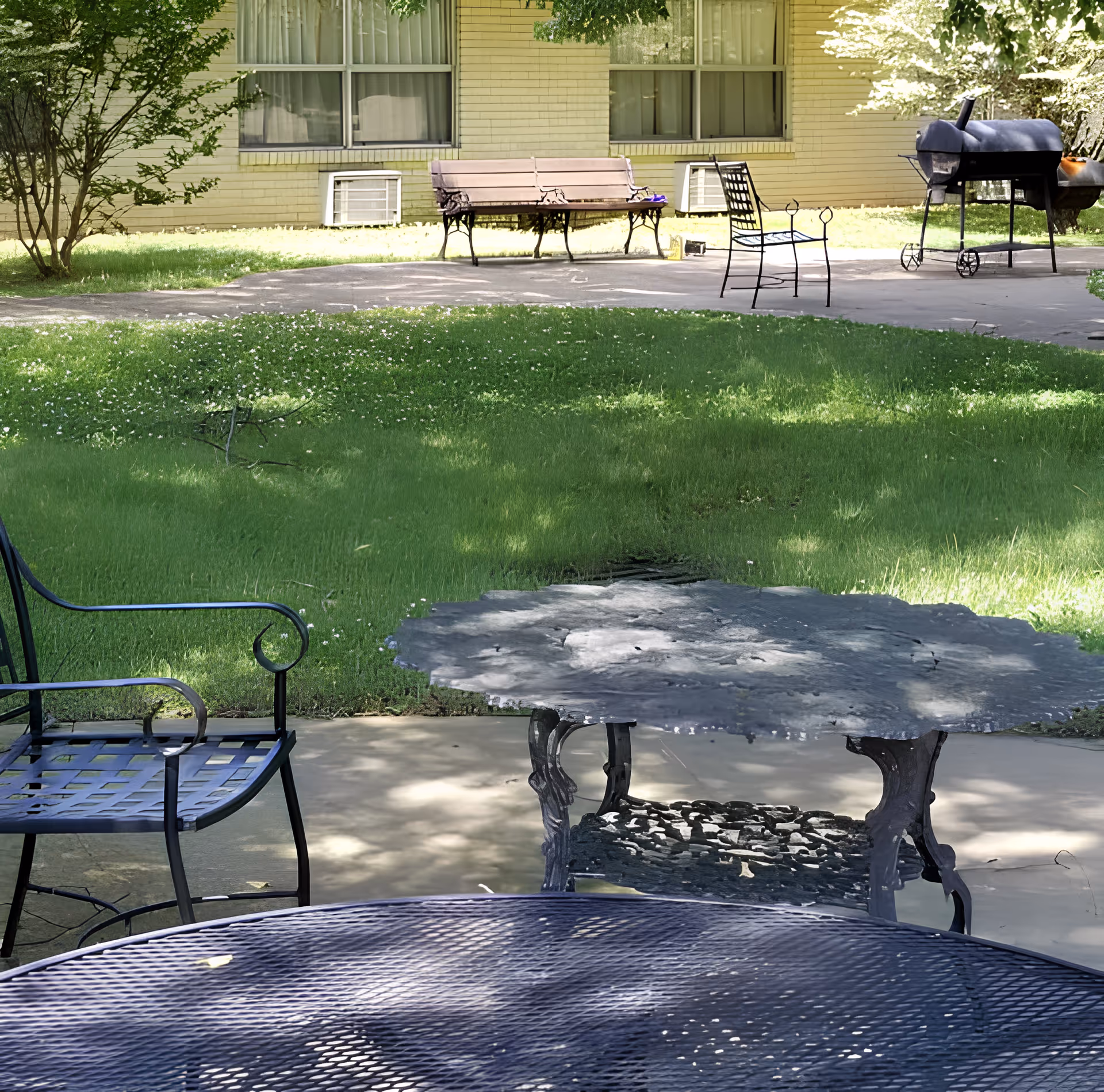 Shaded outdoor courtyard with metal tables and chairs, a bench, grill, and lawn in front of a brick building.