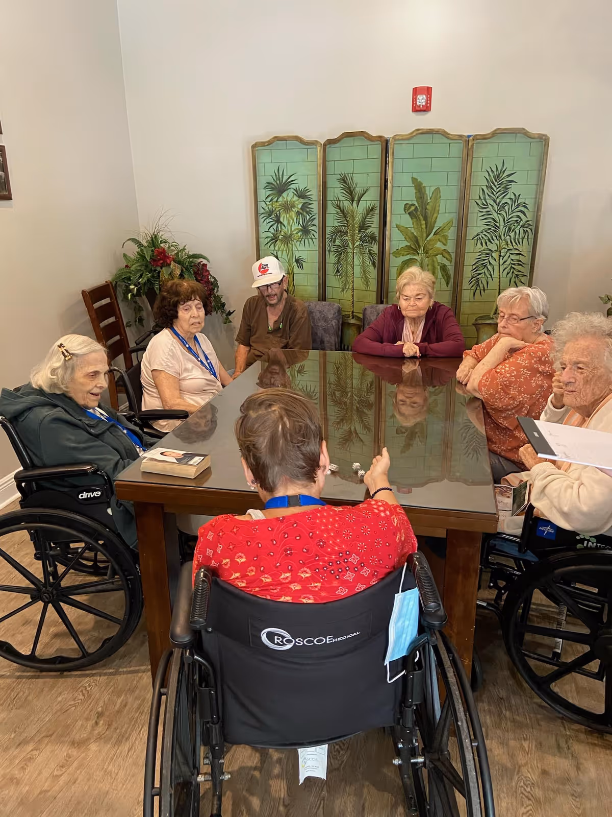 A group of elderly residents, several in wheelchairs, seated around a large glass-topped table in a communal room with a decorative folding screen behind them.