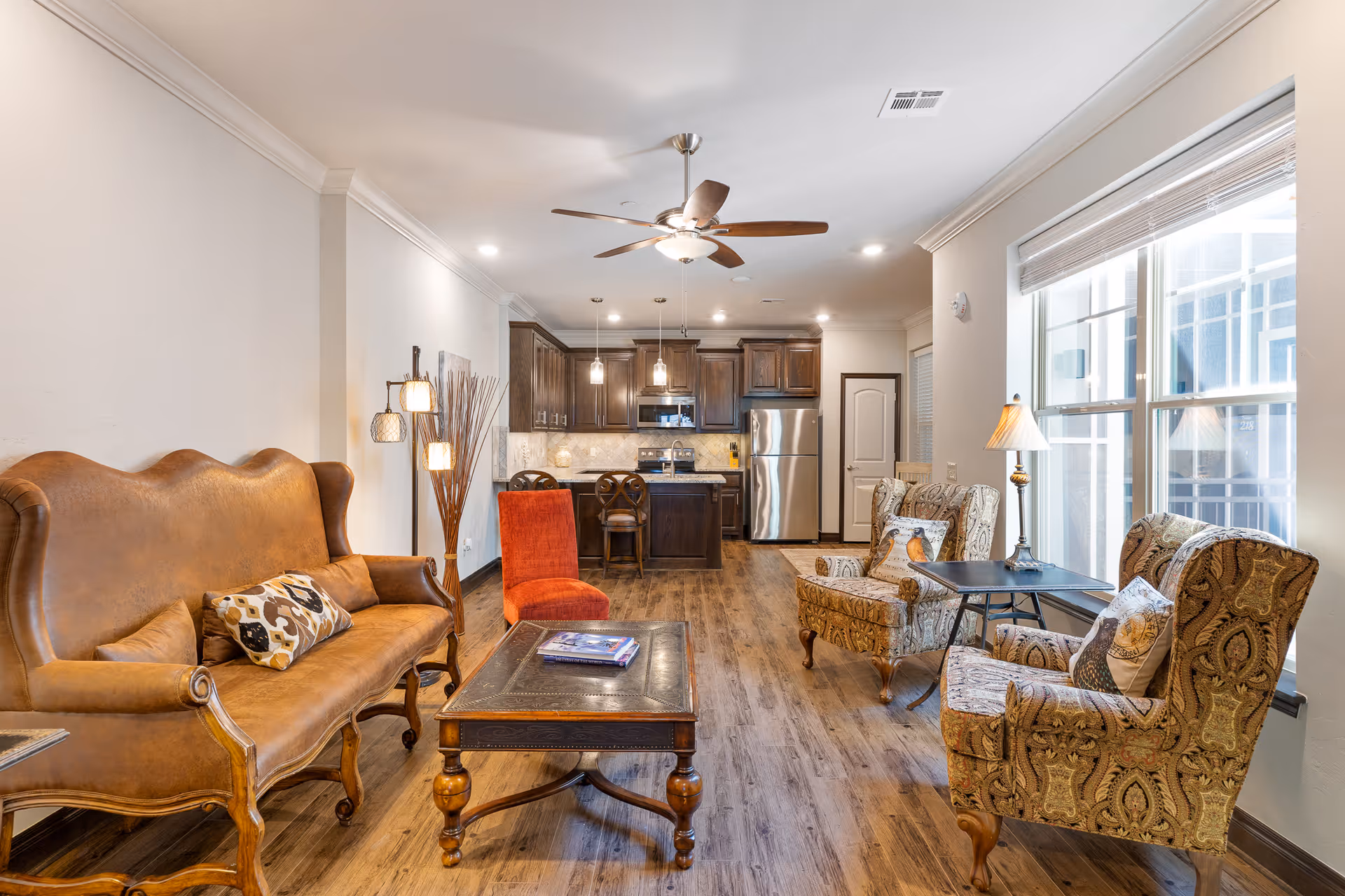 A cozy living room and kitchen area with wooden flooring. The living room features a brown leather sofa with a patterned pillow, two patterned armchairs with pillows, a wooden coffee table with books, and a small side table with a lamp. The kitchen in the background has dark wooden cabinets, stainless steel appliances, and a kitchen island with two chairs. A ceiling fan with lights is mounted on the ceiling, and large windows allow natural light to fill the space.