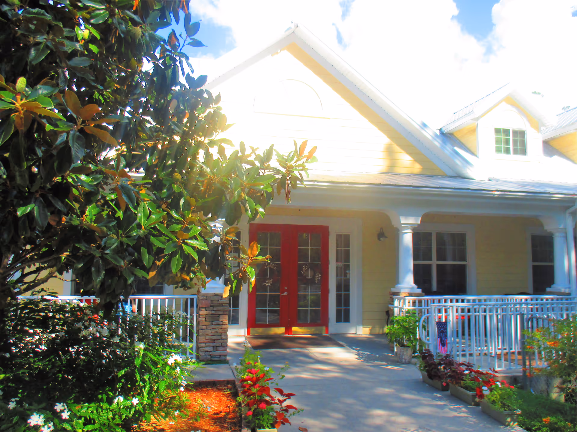 Front entrance of a yellow building with a red double door, white porch columns and railings, and surrounding plants.