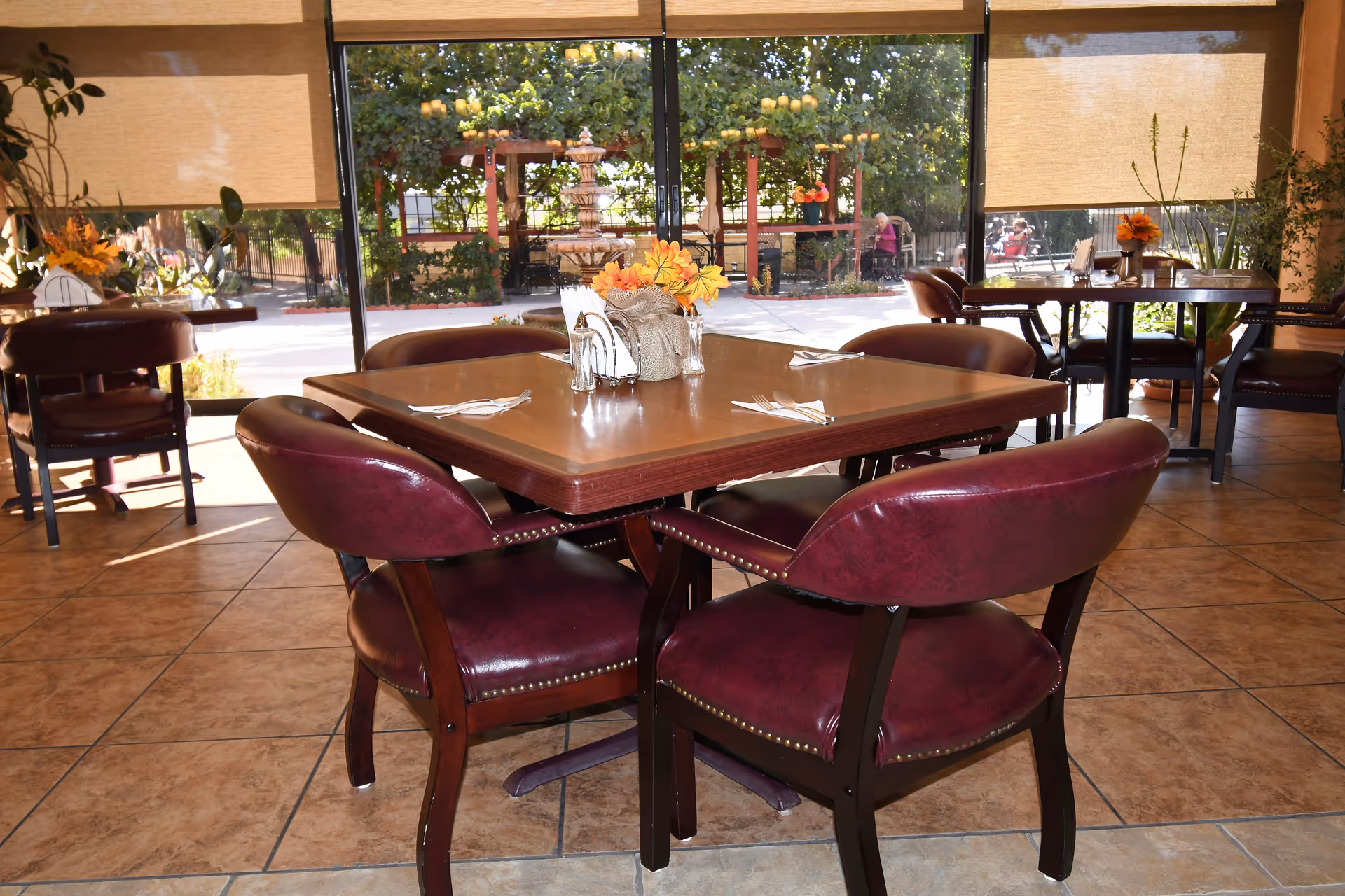 Dining area with a square wooden table set with napkins and silverware, surrounded by four maroon cushioned chairs. The room has large windows with beige blinds partially drawn, allowing a view of an outdoor garden area with a fountain and greenery. Additional tables and chairs are visible in the background, along with some potted plants and floral centerpieces on the tables.