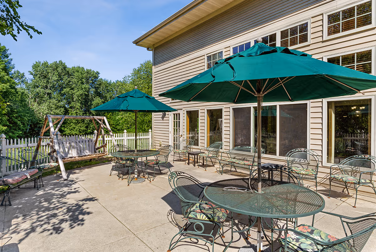 Outdoor patio area with metal tables, chairs and green umbrellas beside a beige building and white picket fence.