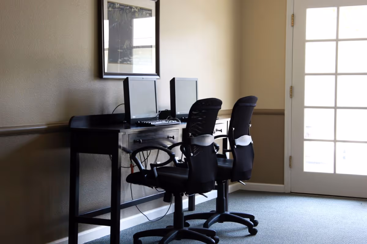 Two black office chairs in front of a dark wooden desk with two computer monitors and keyboards, placed against a beige wall with a framed picture above. A glass door with multiple panes is visible to the right, letting in natural light.