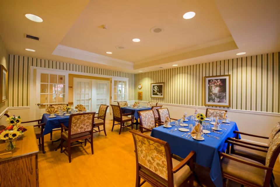 A warmly lit dining room with tables set with blue tablecloths, folded napkins, place settings and floral centerpieces surrounded by upholstered chairs.