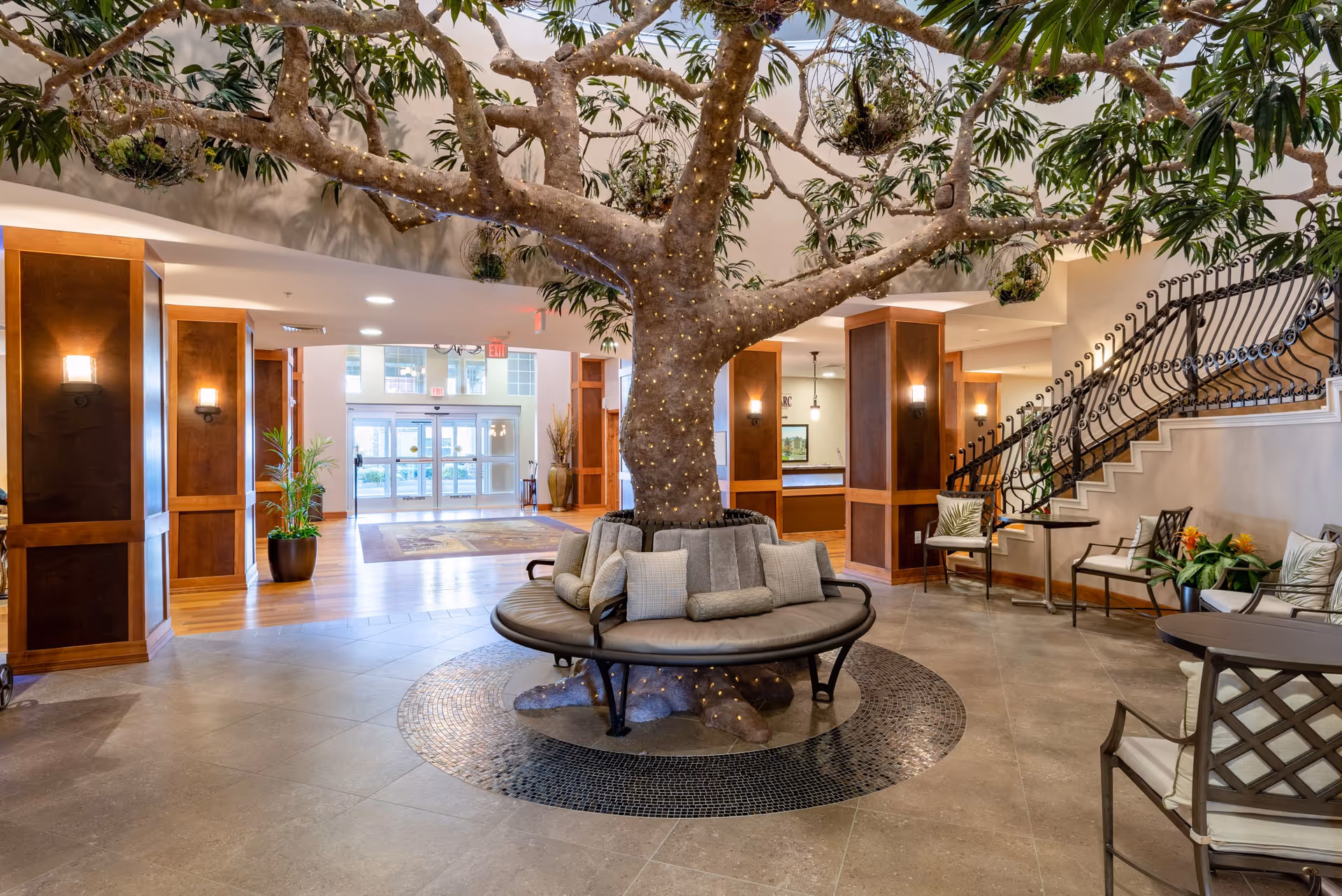 Spacious senior living community lobby featuring a large artificial tree with string lights wrapped around its trunk and branches, surrounded by a circular cushioned bench with pillows. The lobby has tiled flooring, wooden columns with wall sconces, potted plants, a staircase with decorative railing, and seating areas with chairs and tables. Large glass doors are visible in the background providing natural light.