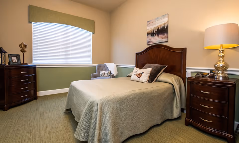 A cozy bedroom with a bed covered in a green bedspread, two decorative pillows, a wooden headboard, a nightstand with a lamp and a phone, a wooden dresser with decorative items, a window with blinds and a green valance, and a gray armchair with a pillow and a white throw.