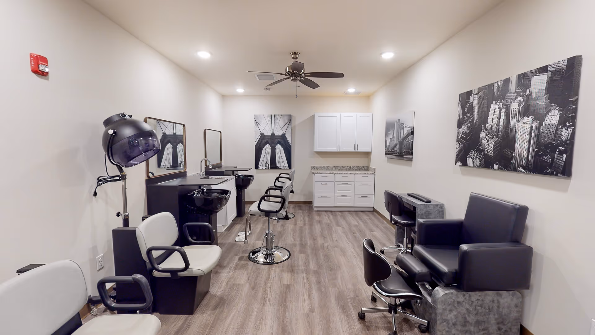 Interior view of a senior living facility's salon area featuring salon chairs, hair washing stations, a hair dryer, and a pedicure chair. The room has light-colored walls, wood flooring, ceiling lights, and black and white cityscape artwork on the walls.