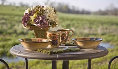 A round outdoor table set with two bowls, two mugs, a small bouquet of flowers in a vase, and some green herbs, with a blurred grassy field in the background.