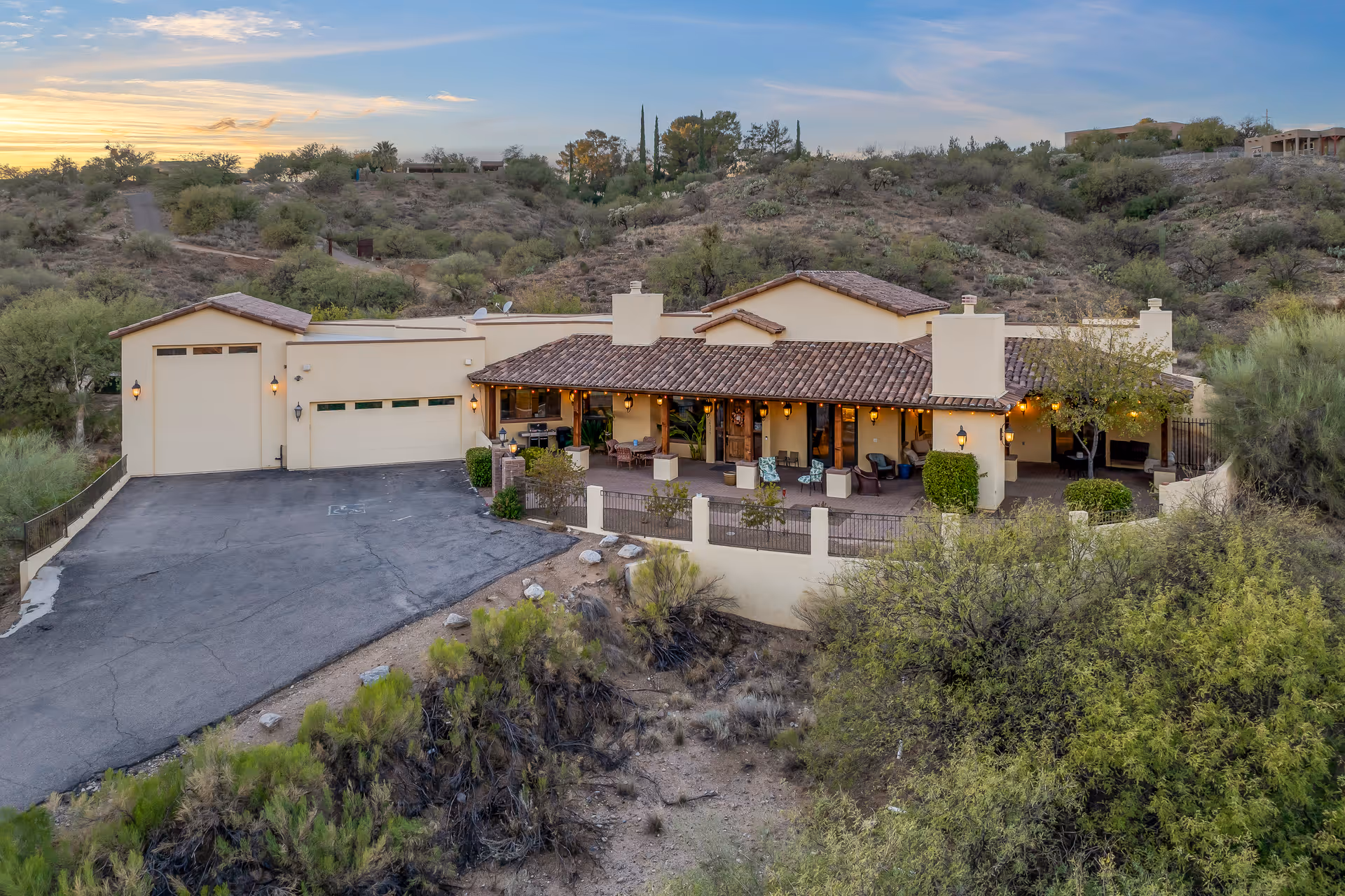 Exterior view of a single-story assisted living facility named Heritage Manor Assisted Living, featuring a tiled roof, a spacious covered patio with outdoor seating, surrounded by desert landscaping and hills in the background during sunset.