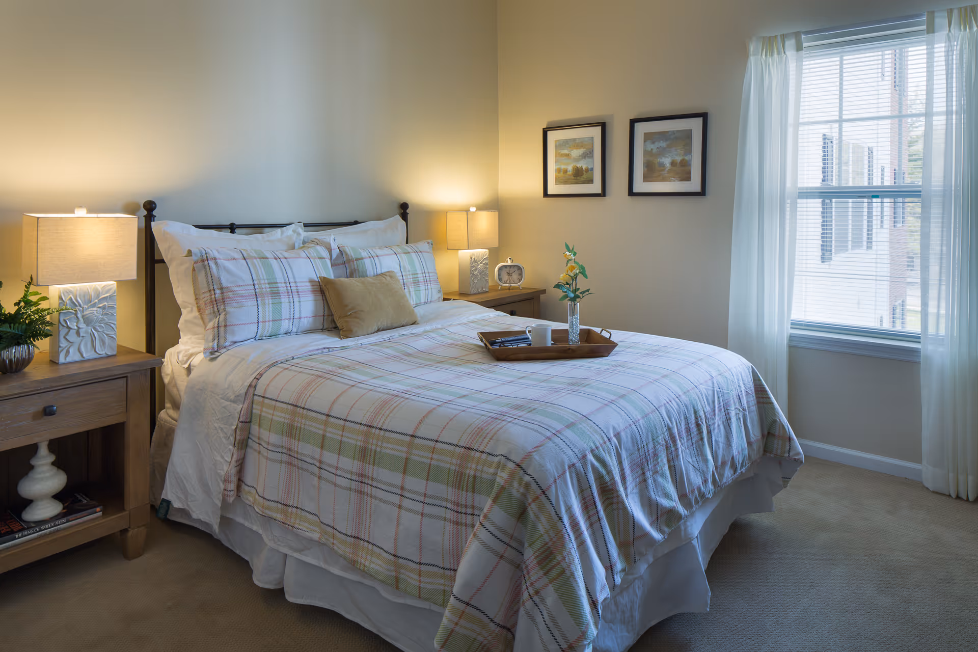 A cozy bedroom with a neatly made bed featuring plaid bedding and multiple pillows. There are two wooden nightstands on either side of the bed, each with a lamp and decorative items. A window with sheer white curtains allows natural light to brighten the room. Two framed landscape paintings hang on the wall above the nightstand on the right.