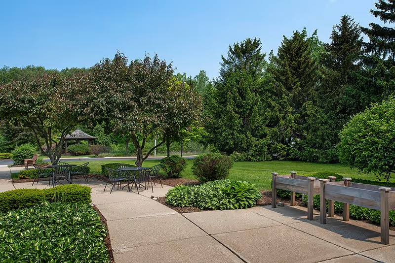 Outdoor garden area with paved walkways, green shrubs, trees, and several black metal tables and chairs. There is a wooden raised garden bed on the right side and a small gazebo visible in the background under a clear blue sky.
