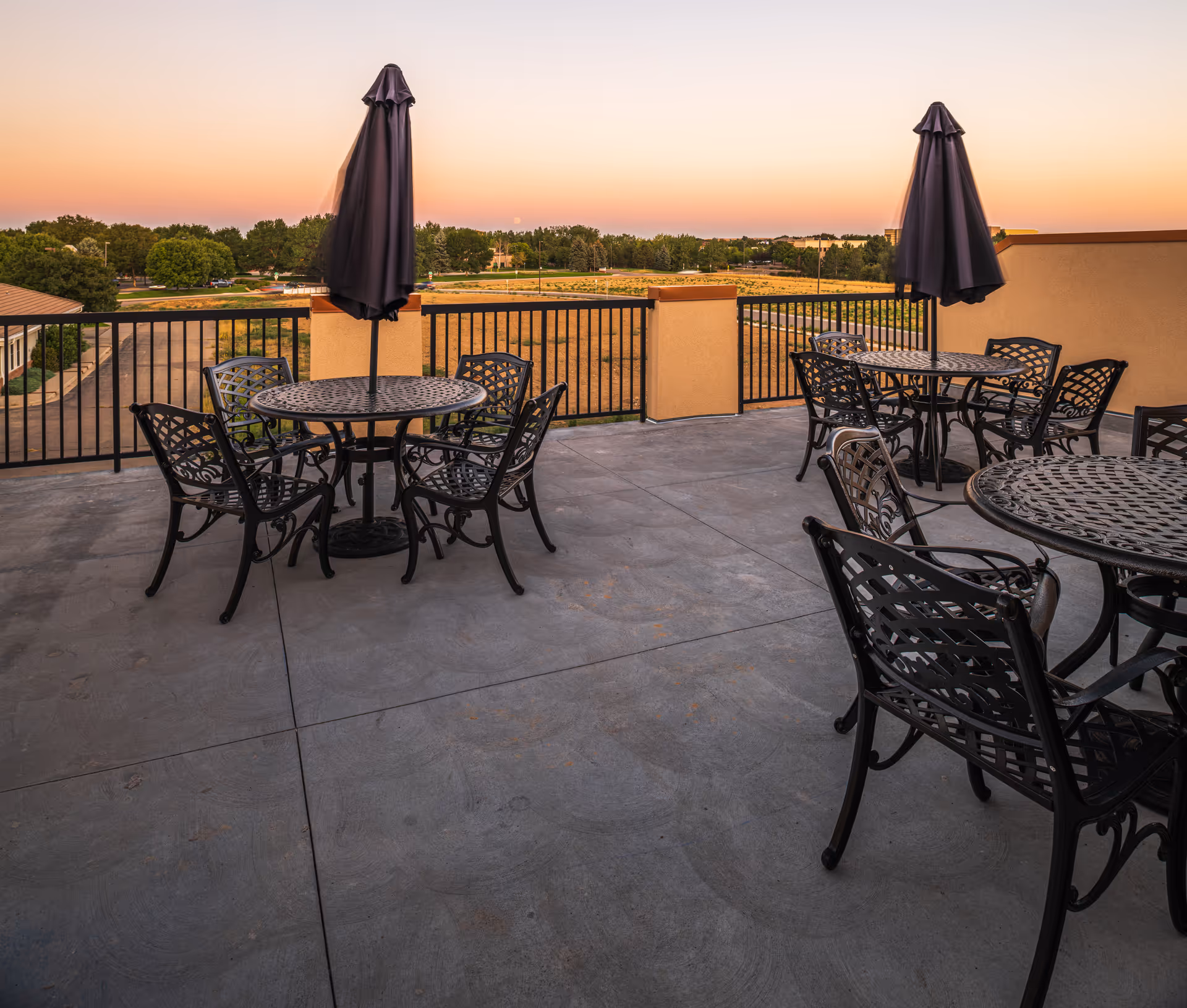 Rooftop patio with metal tables, chairs, and closed umbrellas overlooking a landscaped field at sunset.