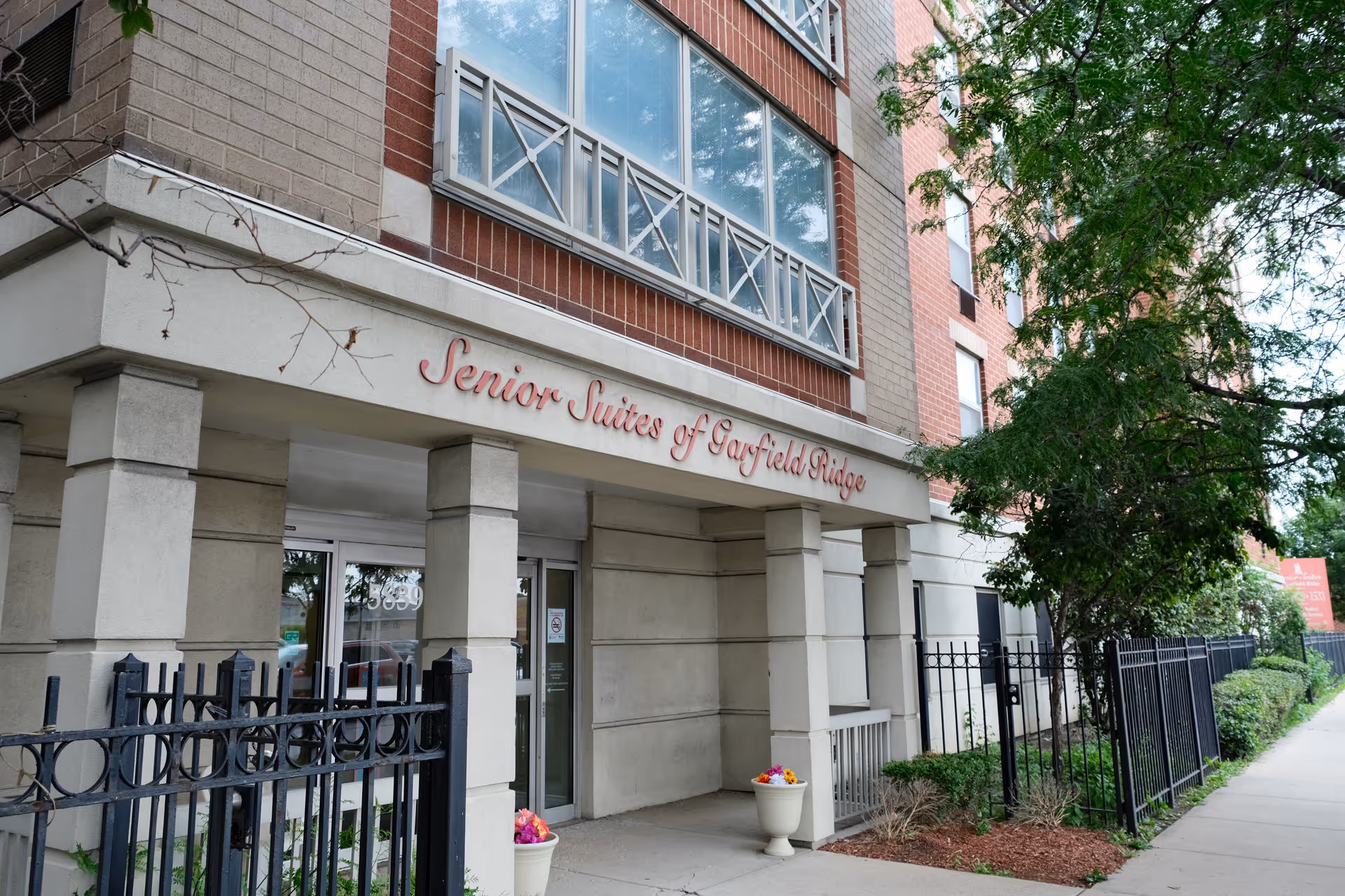 Exterior front entrance of Senior Suites of Garfield Ridge building with a covered entryway supported by columns, a black metal fence, potted flowers, and trees along the sidewalk.