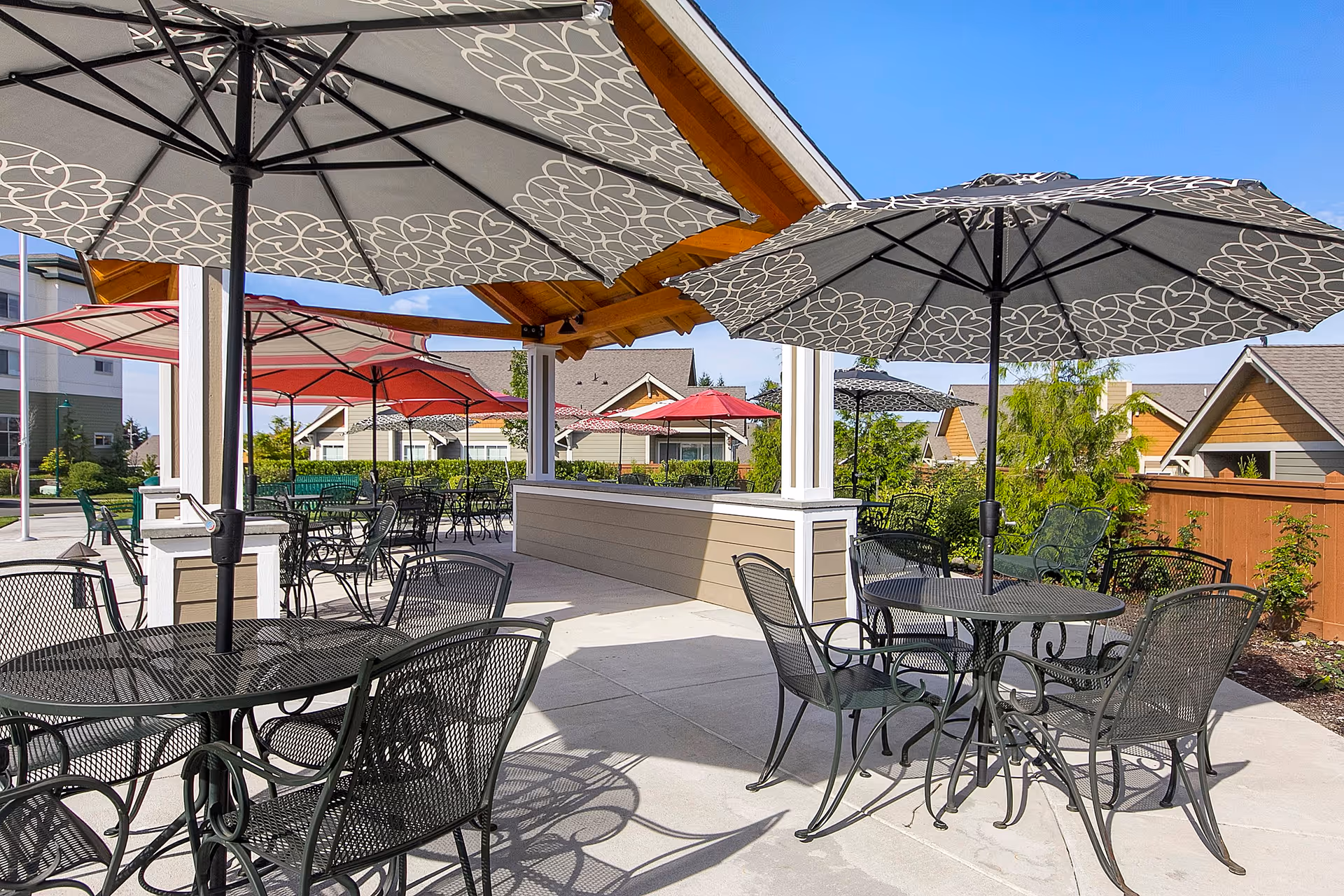 Outdoor patio area with multiple black metal tables and chairs, each shaded by large umbrellas with patterned fabric. The patio is surrounded by a wooden fence and nearby residential-style buildings are visible under a clear blue sky.