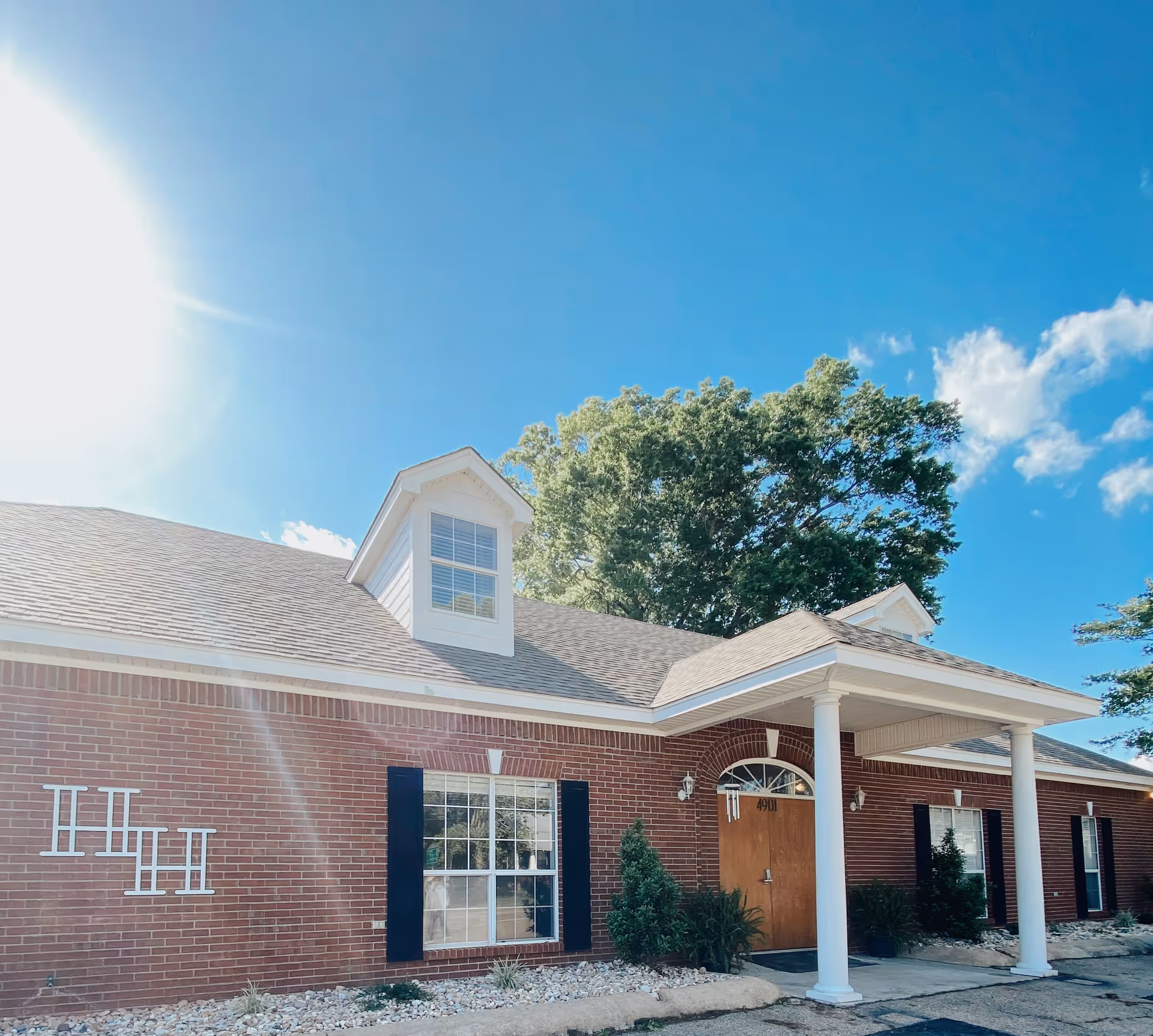 Exterior view of a single-story brick building with white columns at the entrance, a wooden door, multiple windows with black shutters, and a clear blue sky with some clouds and sunlight.
