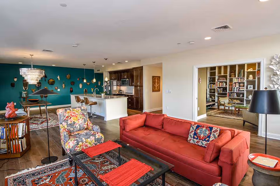 Open living area with a red sofa, floral armchair, coffee table, and view into a kitchen and adjoining library with a teal accent wall.