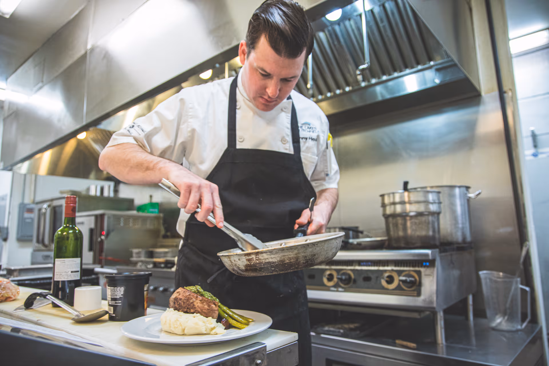 A chef in a commercial kitchen plates a meal of meat, mashed potatoes, and asparagus.