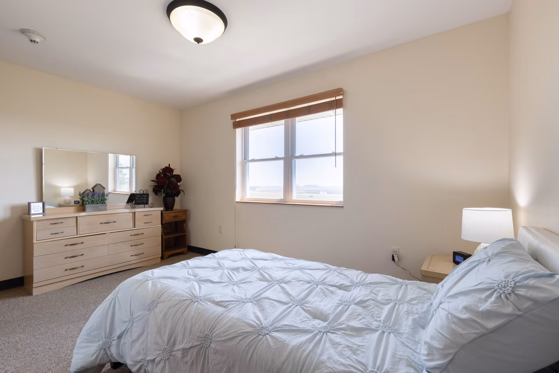 Sunlit furnished bedroom with a made bed, dresser, nightstand and a window.