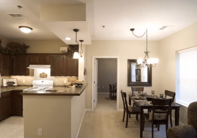 Interior view of a senior living community showing a kitchen area with dark wooden cabinets, a white stove, and a countertop with pendant lights. Adjacent to the kitchen is a dining area with a dark wooden table set for four, a chandelier above, and a large window with blinds. A doorway leads to another room in the background.