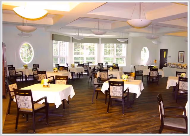 A bright dining room with multiple tables covered in white tablecloths, each surrounded by dark wooden chairs with patterned cushions. The room has large windows letting in natural light, circular windows on the side walls, and several hanging light fixtures from the ceiling. There are small flower arrangements on each table and a buffet area in the back corner.