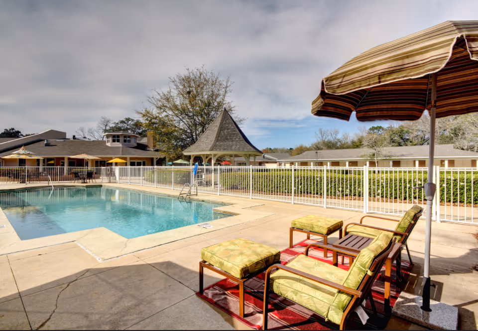 Outdoor swimming pool area with lounge chairs and umbrellas. The pool is surrounded by a white fence, and there is a gazebo and buildings in the background under a partly cloudy sky.